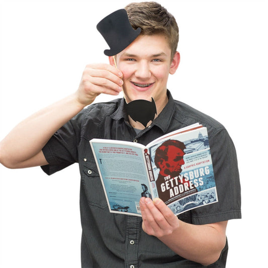 Boy holding The Gettysburg Address open in left hand while holding a black paper top-hat and beard with his right hand with a white background.